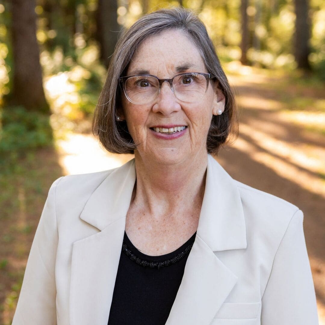 Debbie Christiansen Stowe, VP Quebec, CFUW Portrait of Debbie Christiansen Stowe standing on a wooded path, wearing a light-coloured blazer and glasses, with trees and soft natural light in the background.