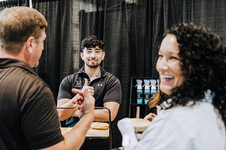 Image of a business employee looking and smiling at the camera with a thumbs up, while other people are laughing and chatting.