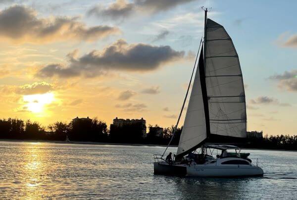 Photo of catamaran sailing along on the water with a sunset backdrop.