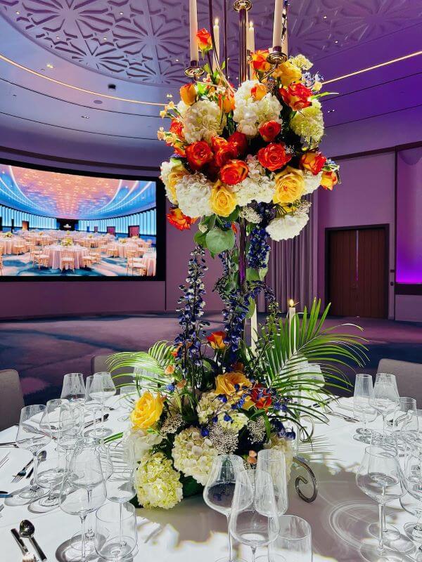 Picture of a tall floral arrangement on top of a dining table in a large ballroom. The floral arrangement has white, red, yellow, and purple flowers.