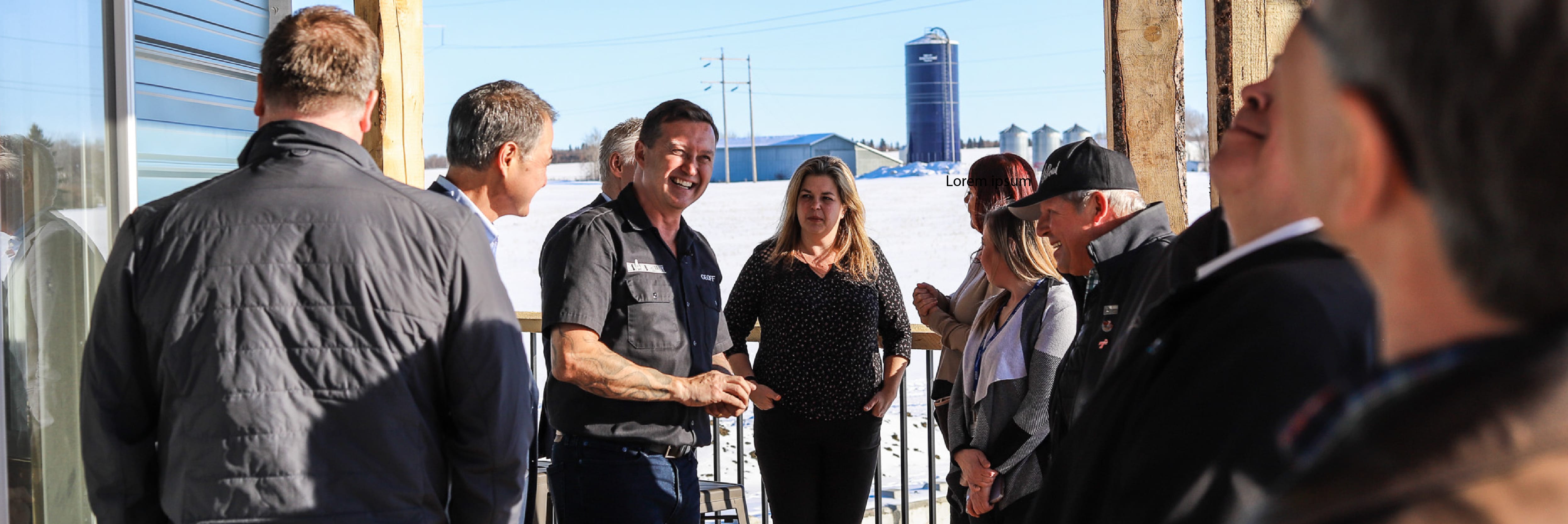 Group of community members gather around business owner on an outdoor deck. The business owner smiles amid telling a story and the audience is engaged, silos and fields can be seen off the deck.