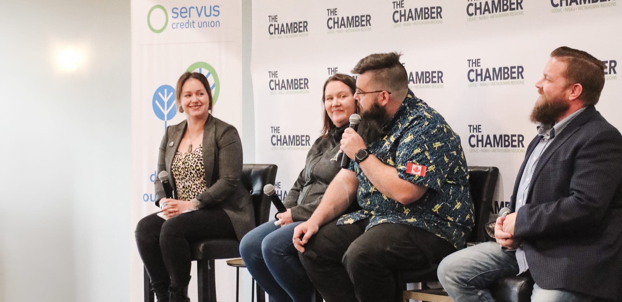 A panel of four speakers smile in front of a Chamber backdrop, they sit on tall stools.