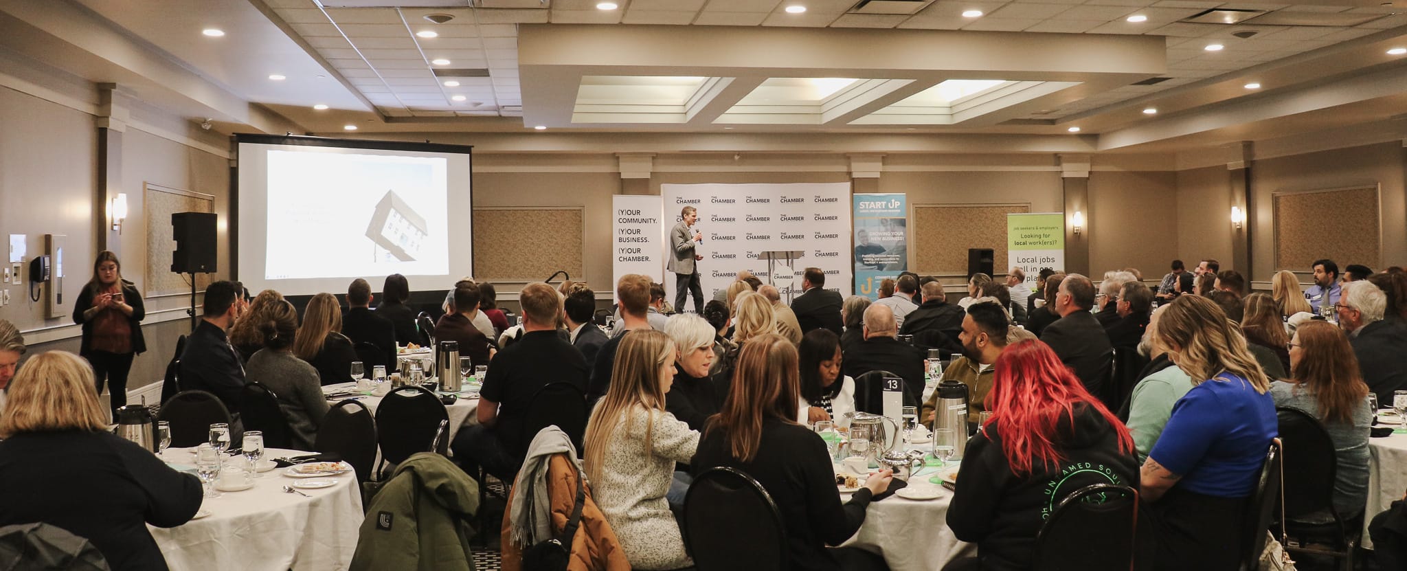 Wide image of a room full of people, a large screen has a pie chart on it and one speaker is on the stage.