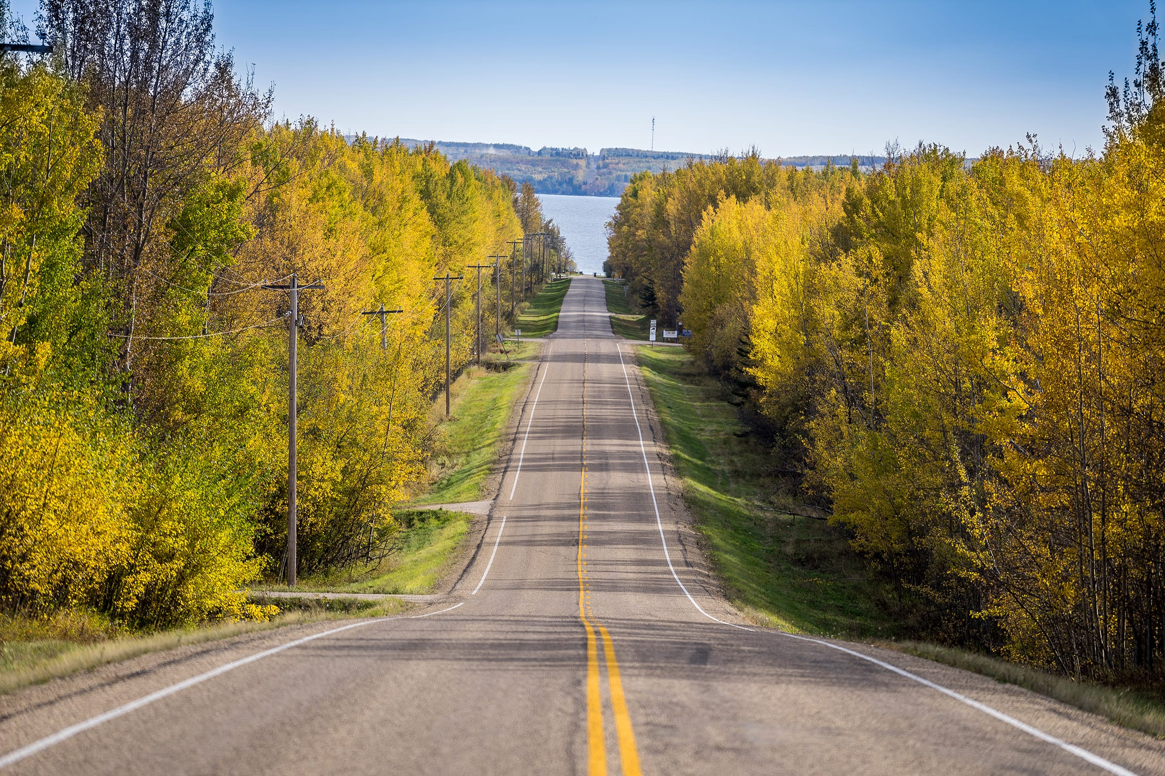 Bright fall image of a single lane highway leading to a lake in the distance.