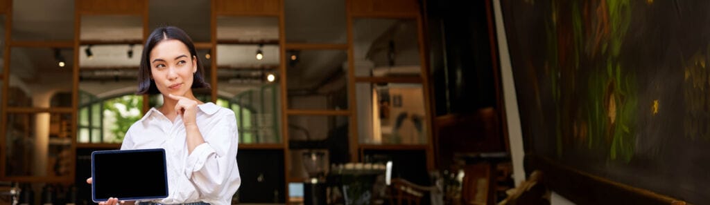 Young business owner Young woman looks thoughtful, stands in cafe, shows her digital tablet screen.