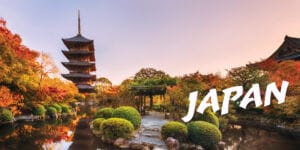 A serene japanese garden with shrubery, trees with blossoms and a temple overlooking a serene pond.