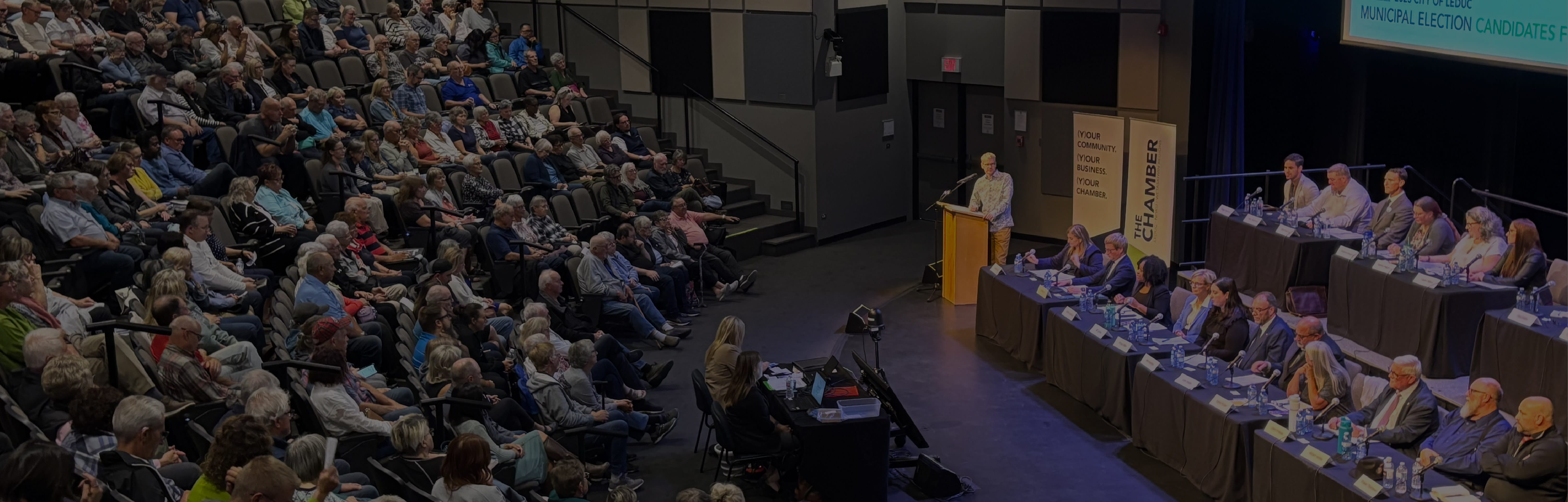 The leduc municipal election candidates forum as seen from above. The candidates are on the stage the moderator looks towards them from his podium and the crowd watches attentively.