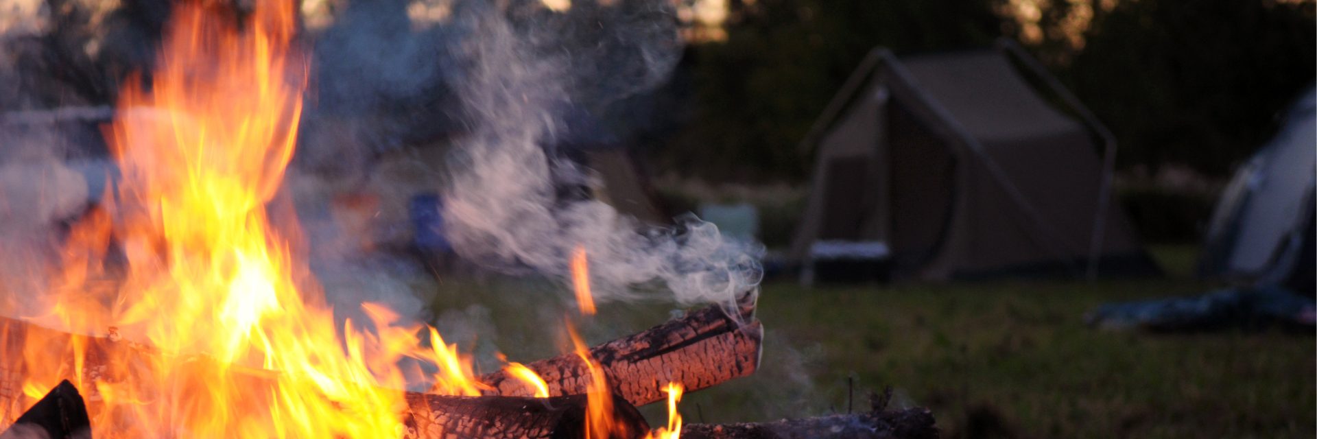 Campfire burning in the foreground with a blurred tent in the background at a campsite during dusk.