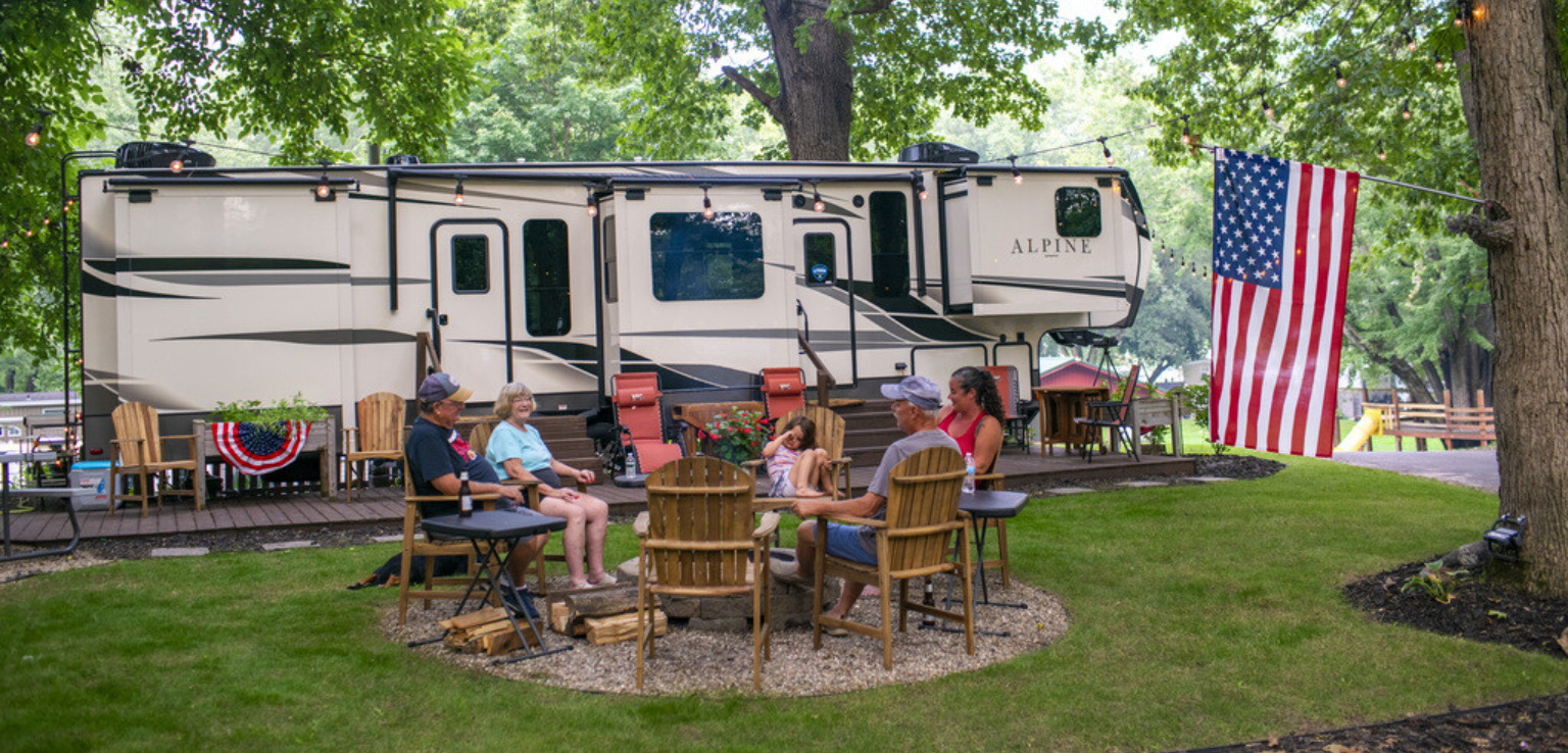 A diverse group of individuals relaxing around a fire pit, with an RV parked nearby, creating a warm, inviting atmosphere.
