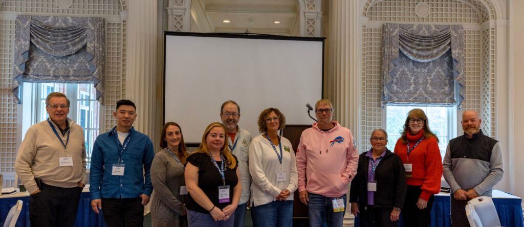 CONY 2025 Meeting_24 A group of eleven people stand indoors in front of a projector screen, posing for a photo, all wearing conference name badges.