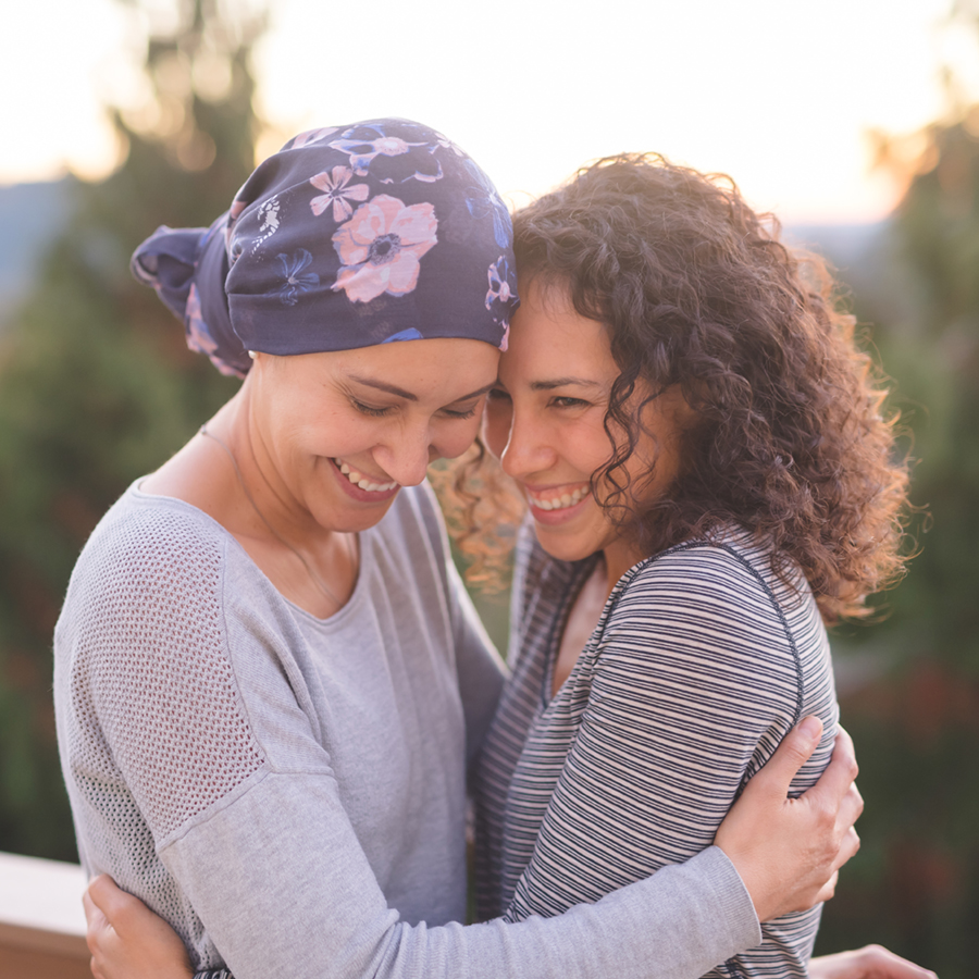 A beautiful young ethnic woman fighting cancer and wearing a head wrap embraces her sister. They are tightly holding each other and she is looking down and smiling. Her sister is also smiling. They are standing outdoors and there are mountains and trees in the background.