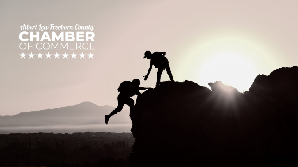 a person gives a hand to another climbing a mountain. The Albert Lea-Freeborn County Chamber logo is in the top left corner