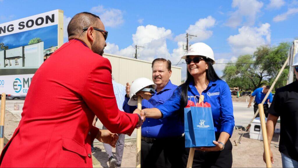 A woman in a hard hat and blue shirt is receiving a gold shovel and a blue gift bag from a man in a red jacket
