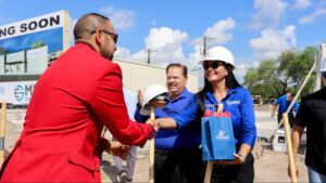 A woman in a hard hat and blue shirt is receiving a gold shovel and a blue gift bag from a man in a red jacket