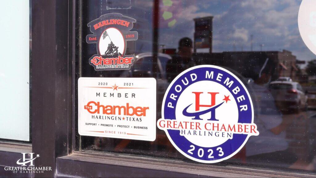 A glass door with multiple stickers on it, including "Proud Member of Greater Harlingen Chamber 2023" and "Member of The Chamber Harlingen, Texas Since 1919."