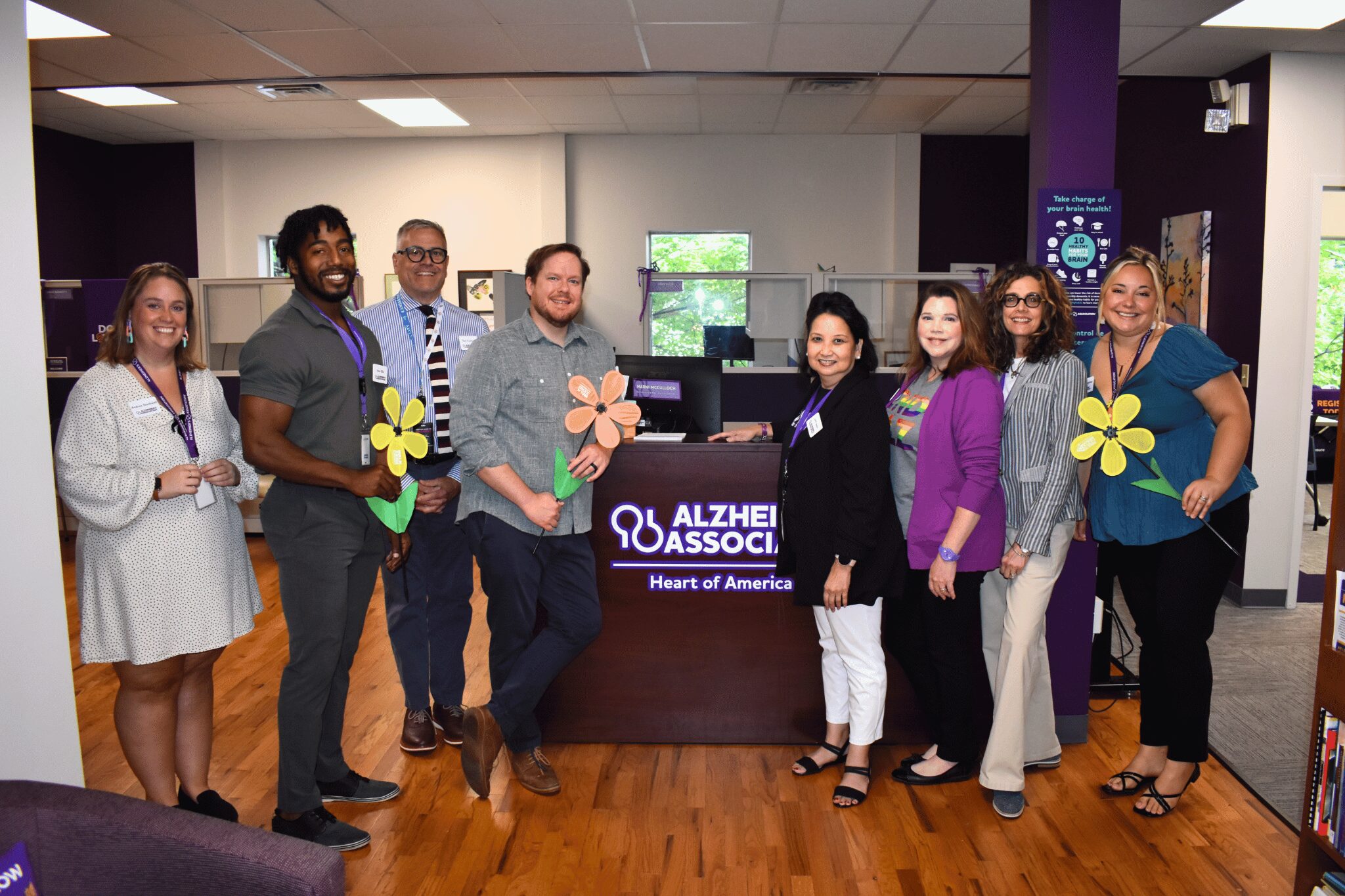 Coffee Connections Aug Alzheimers Association The Alzheimer's Association team stands in front of their logo in their office space, posing for the picture.