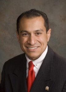 Portrait of Corey Calaycay wearing a black suit, white shirt, and red tie, smiling against a neutral background