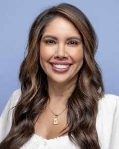 A smiling woman with long, wavy hair, wearing a white top and a necklace, against a light blue background.