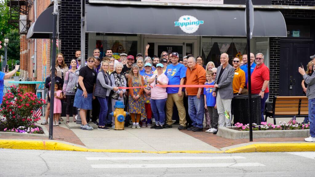 A photo of the ribbon cutting ceremony outside the entrance of Jamie's Cafe in Bluffton, IN. The group cutting the ribbon consists of Jamie's Cafe staff/employees, Chamber staff, Chamber ambassadors, and local community members.
