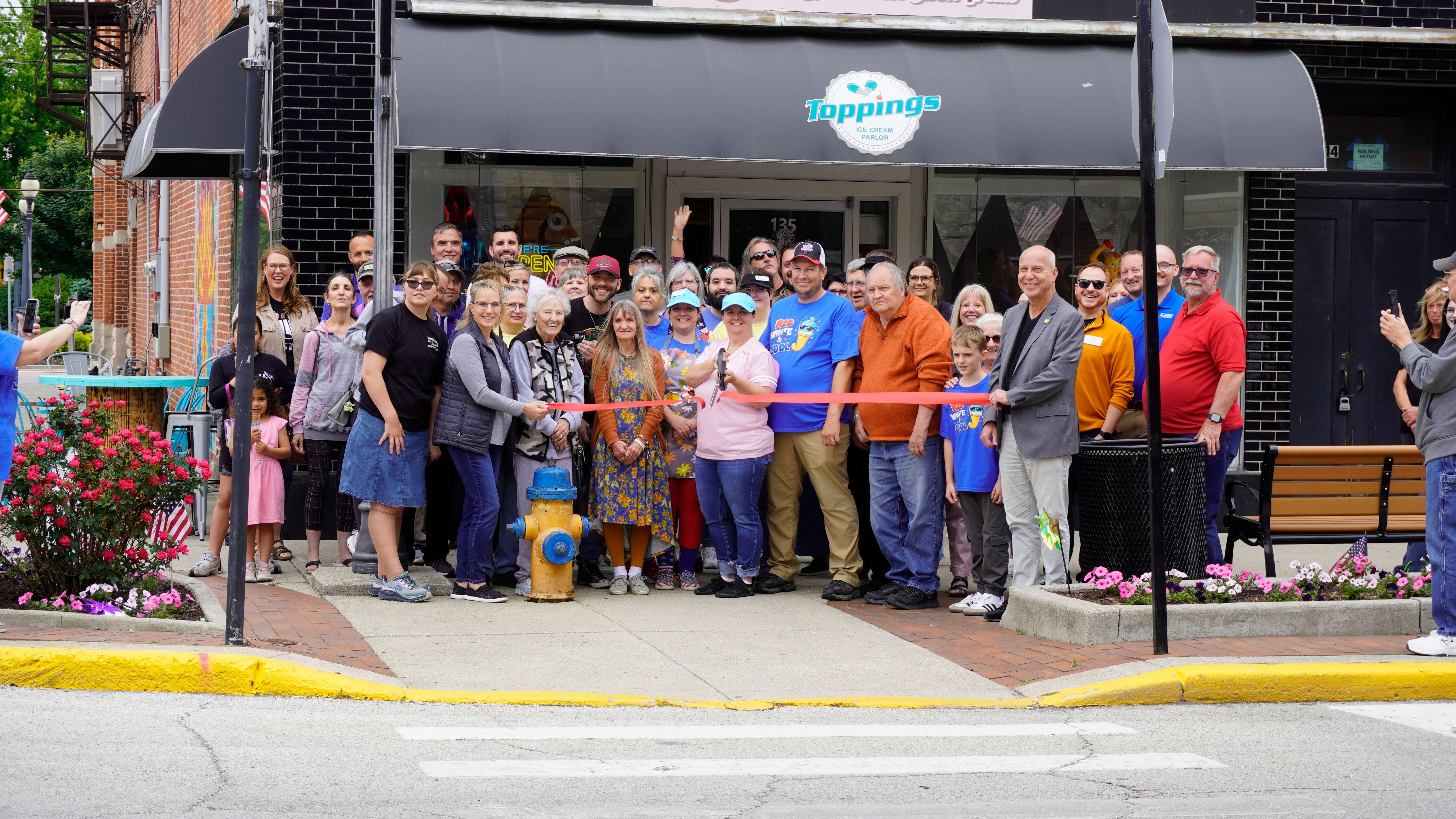 A photo of the ribbon cutting ceremony outside the entrance of Jamie's Cafe in Bluffton, IN. The group cutting the ribbon consists of Jamie's Cafe staff/employees, Chamber staff, Chamber ambassadors, and local community members.