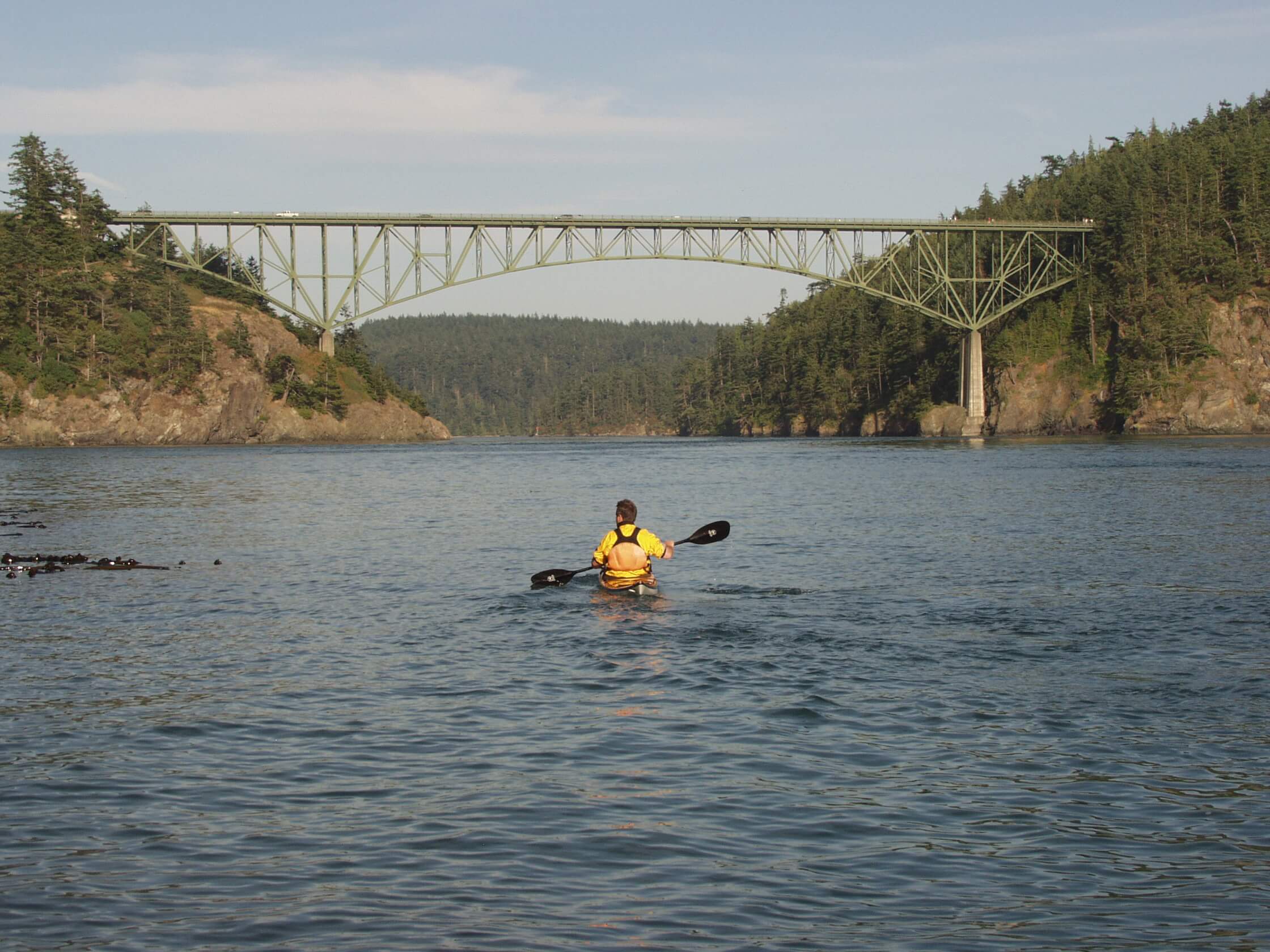 A kayaker wearing a yellow life jacket paddles across calm water toward a large green steel arch bridge spanning between two forested cliffs under a cloudy sky.