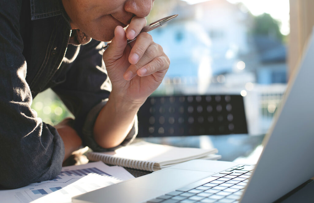 Pensive businessman in smart casual wear looking at laptop computer screen during working in modern office, planning and thinking about new project, analyzing market report, financial document on table, close up. Business planning and strategy analysis concept