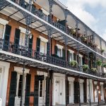 New Orleans architecture photo shows brick 3-story building, black shutters, white trim, ornate, black wrought iron railings, hanging green ferns, and green flower baskets on railings