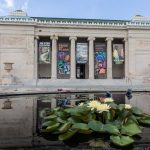 New Orleans Museum of Art displaying a vertical banner between each of its columns. Lily pads in a water fountain are seen in the foreground.