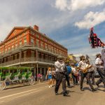 Second line marching in French Quarter with horse-drawn carriage parked and three-story brick building behind second line.