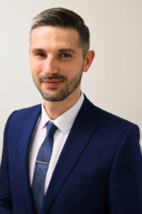 Andy Clare headhost, man with short brown hair, beard, mustache, wearing navy suit, white dress shirt and navy tie