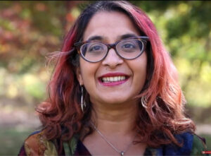 Anu Subramanian headshot, women with black hair, red highlights, dark rimmed classes, wearing multi-colored blouse