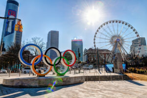 Daytime photo of bright sun shining on Atlanta downtown buildings, ferris wheel and Olympics logo sculpture.