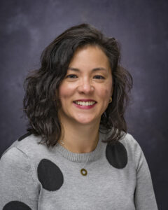 Jennifer Phelan headshot, woman with curly, brown, shoulder-length hair, wearing gray sweater with black dots on it