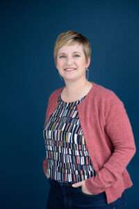 Tammy Fredrickson headshot, woman with short brown hair, highlighted blonde, wearing multicolored blouse, pink cardigan, and black pants