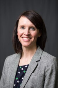 Heather Isaacson headshot, white female, dark brown, shoulder-length hair, Gray suit jacket, black polka dot blouse