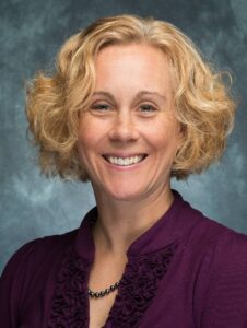 Jennifer Essig headshot, female with short, curly blonde hair, plum-colored blouse