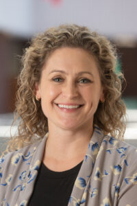 Alli Sauerwein headshot, white female, shoulder-length, curly blonde hair, gray cardigan, black blouse