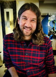 Man with brown wavy hair to just below shoulders wearing red and blue plaid button down shirt