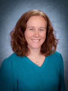 Woman with reddish brown, shoulder-length, wavy hair wearing a teal v-neck top.