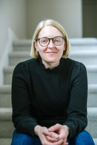 Woman with short, straight blonde hair wearing glasses, black long sleeve top, jeans, and sitting on staircase.