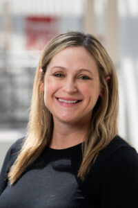 Woman with long blonde-highlighted brown hair, wearing a black top.