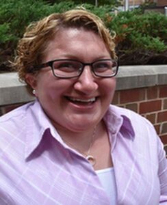 Women with short blonde / brown hair, wearing glasses and a light pink button down shirt.