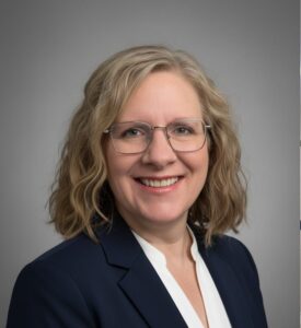 Woman with shoulder-length, wavy, blonde hair wearing glasses, white blouse, and dark blazer