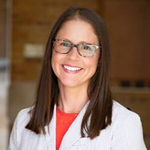 Woman with below the shoulder, straight brown hair, wearing glasses, smiling, wearing a red blouse and white blazer