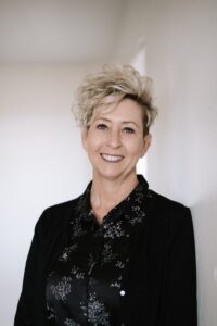 Woman with short highlighted hair, smiling, wearing black floral print blouse and black sweater