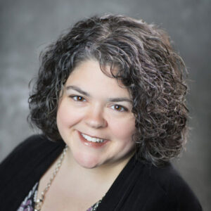 Woman with chin-length, curly, brown hair, smiling, wearing blouse and black sweater