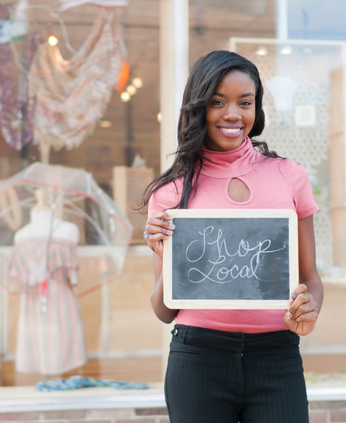 A young black woman standing in front of a clothing boutique with a sign in her hands that reads, "Shop Local."