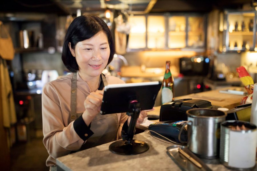 A middle aged asian woman with a slight smile on her face and wearing an apron while interacting with a point of sale system.