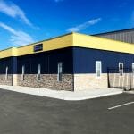 A side view of a one story office building with yellow and blue paint and stone siding in a parking lot