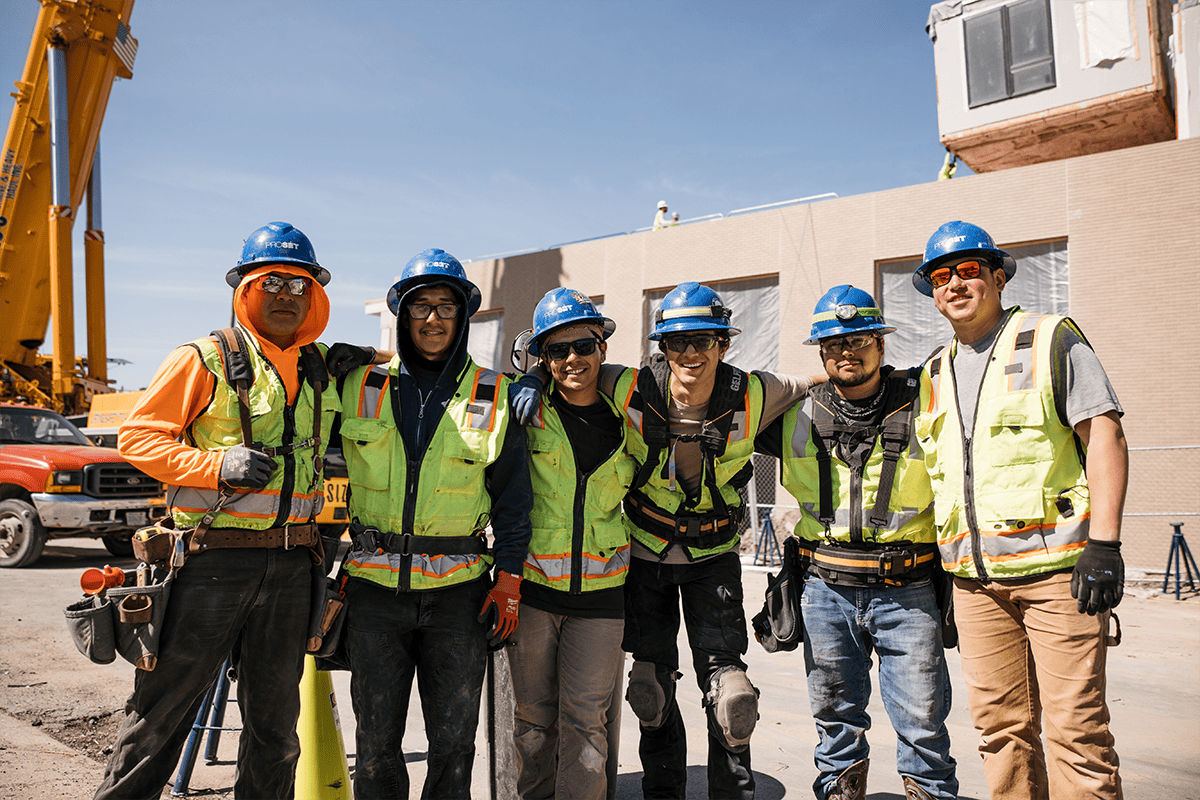 A group of modular building construction workers lean against one another, with their recent modular project in the beginning stages of assembeling behind them